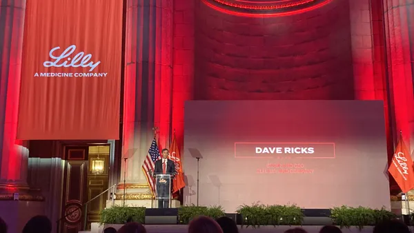 A person speaks from a podium on stage, flanked by red banners.