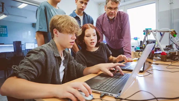 Four  high school students and a teacher are sitting and standing around a desk in a classroom. One student siting at the desk is using a laptop and mouse and others are looking at the screen