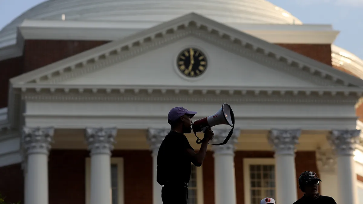 A man standing in front of an academic building shouts using a megaphone a crowd that is just out of frame.