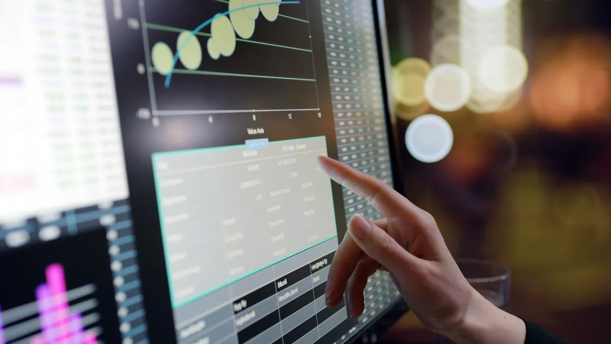 Close-up image of a woman’s hand using a touch screen computer in an office. An array of data is displayed on the screen.