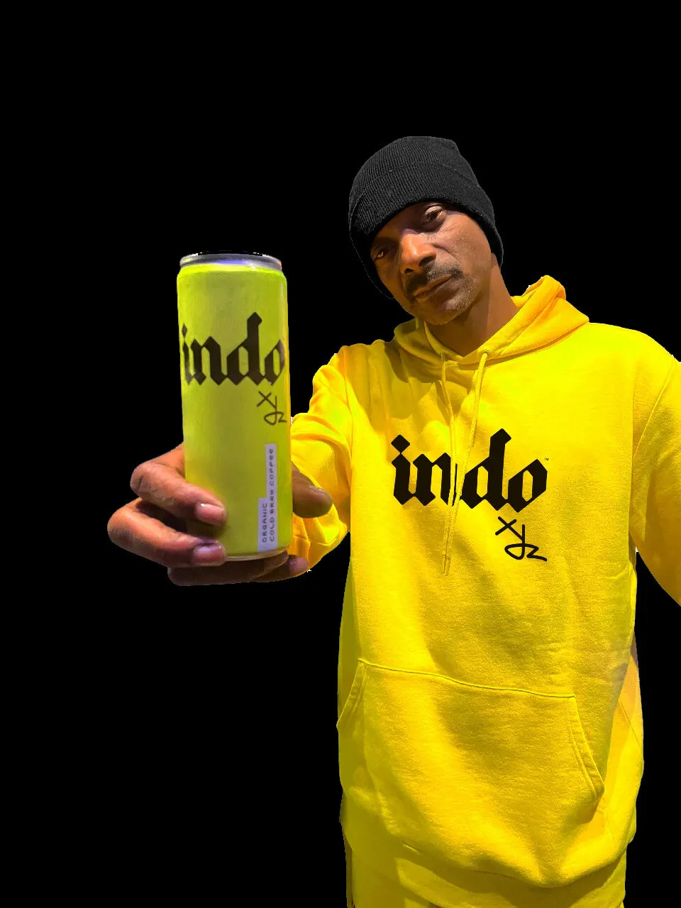 Snoop Dogg stands to the right, wearing a black knit hat and a yellow INDOxyz sweatshirt, holding a yellow can of INDOxyz cold brew toward the camera. The background is black.