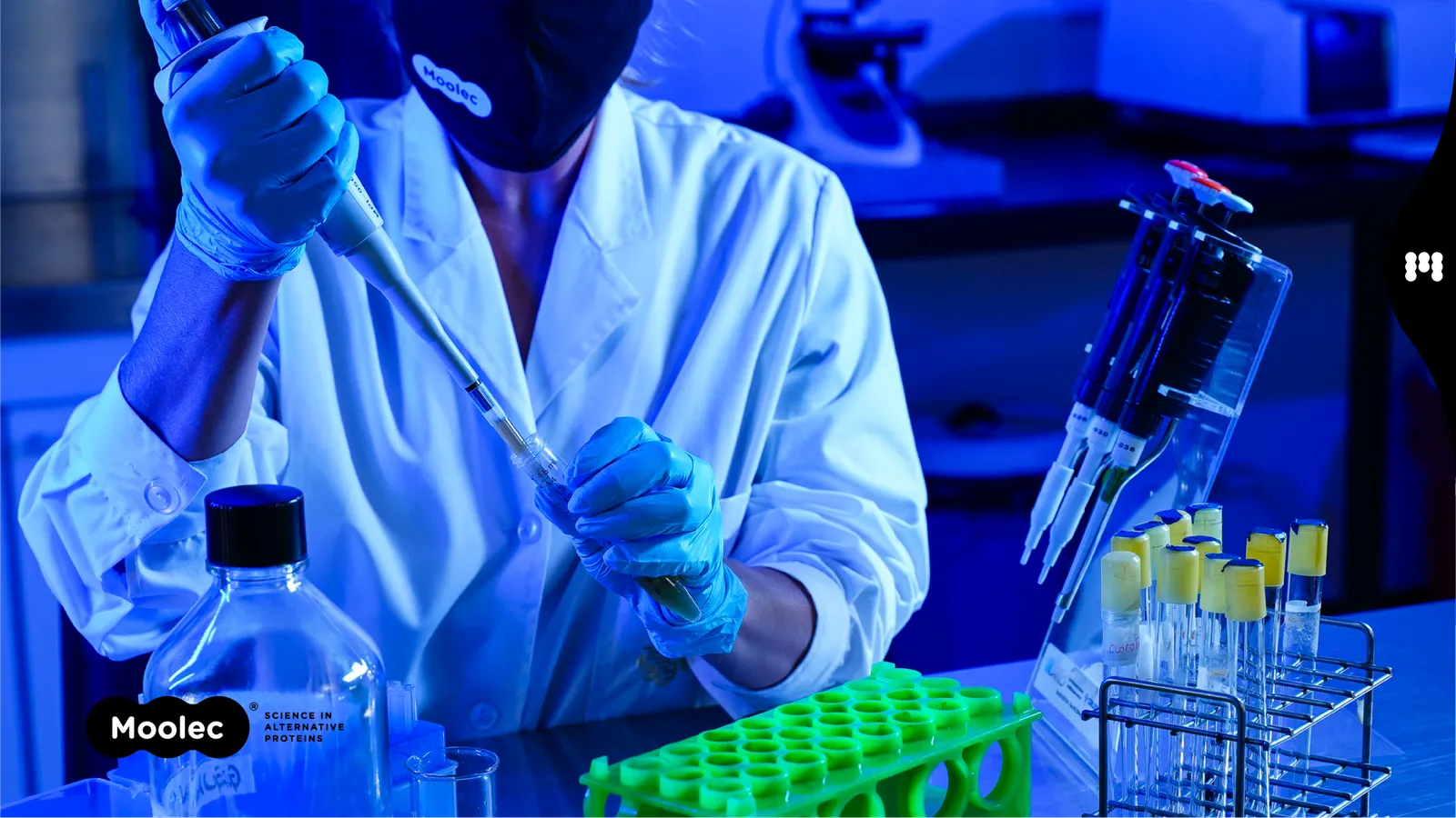 A scientist wearing a face mask, lab coat and rubber gloves puts a substance into a test tube containing a green substance. The person is in a lab with a bluish light, and there is other lab equipment
