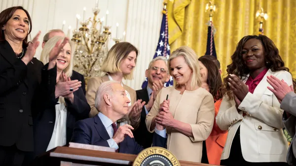 U.S. President Joe Biden hands a pen to former Fox News anchor Gretchen Carlson in the East Room of the White House, surrounded by people.