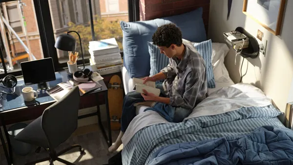A person with short hair and a flannel shirt sits on a blue sheet bed in a small dorm room