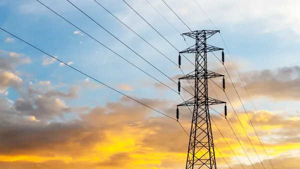 Silhouette of a large electrical transmission tower with power lines against a dramatic sunset sky featuring blue and bright orange clouds.
