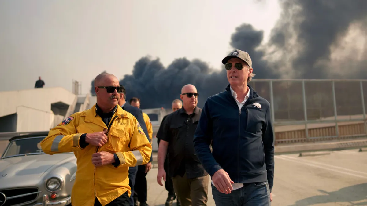 Gov. Newsom walks along a street as smoke is visible behind him.