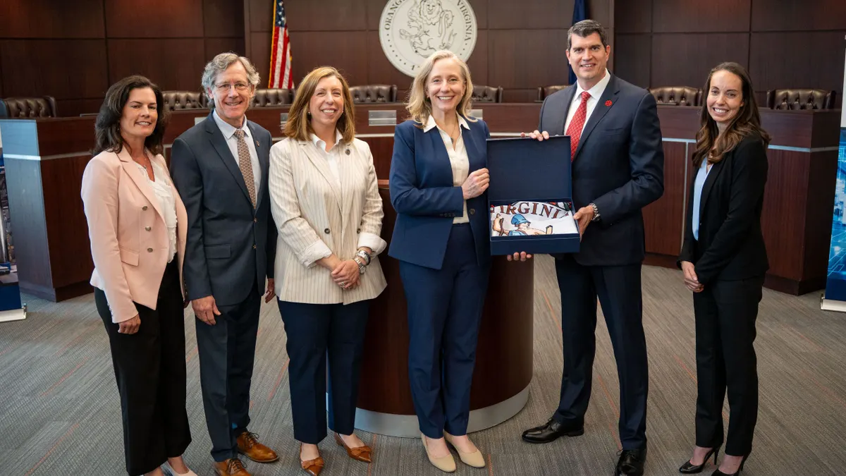 Six people in sports coats and slacks smiling at the camera with dark brown background.