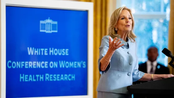 A person in a blue suit speaks at a podium in front of a sign reading "White House Conference on Women's Health Research."