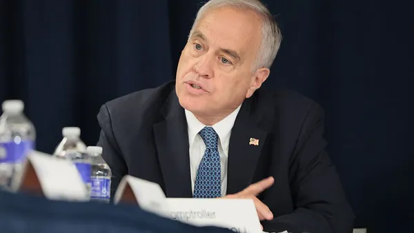 A seated person in a dark suit with a blue tie speaks at a conference table.