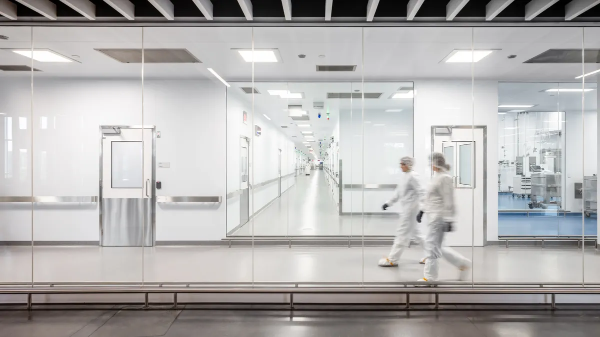 Two individuals in full-body cleanroom suits walk through a sterile, modern pharmaceutical or biotechnology facility