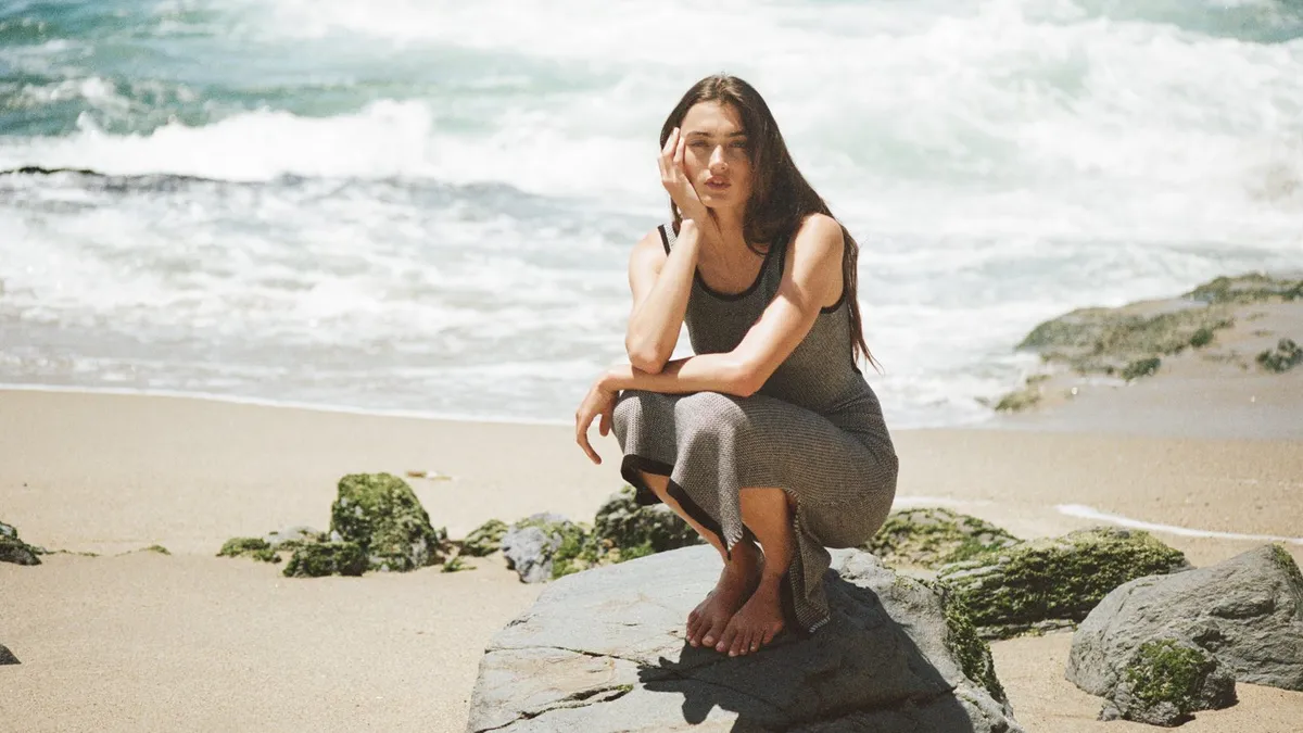 A person with long brown hair in a gray tank dress squats on a rock, on a sandy shoreline, during the day. Her head rests on one hand.