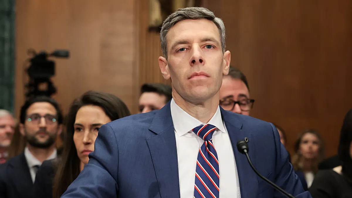 Comptroller of the Currency Jonathan Gould, wearing a suit and tie, sits before a microphone during a Congressional hearing, with people sitting behind him.