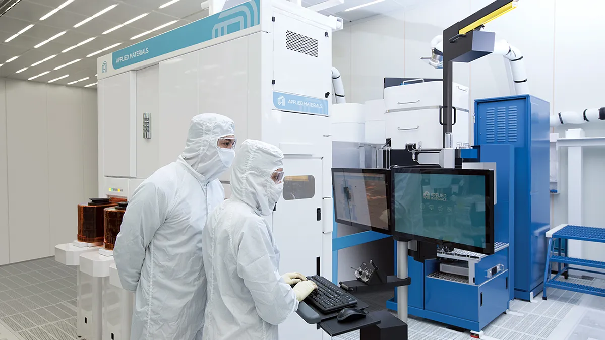 Two employees in PPE work on a computer beside Applied Materials equipment.