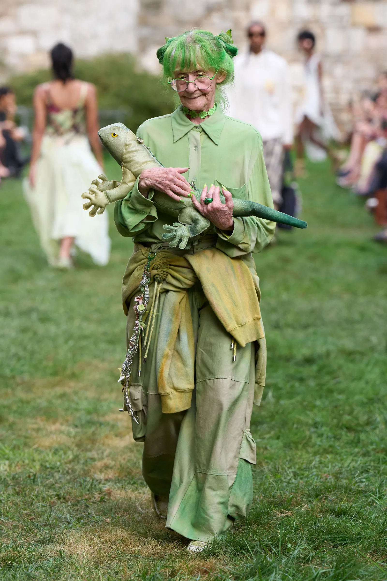 A woman wearing all-green clothing walking on a runway in a garden.