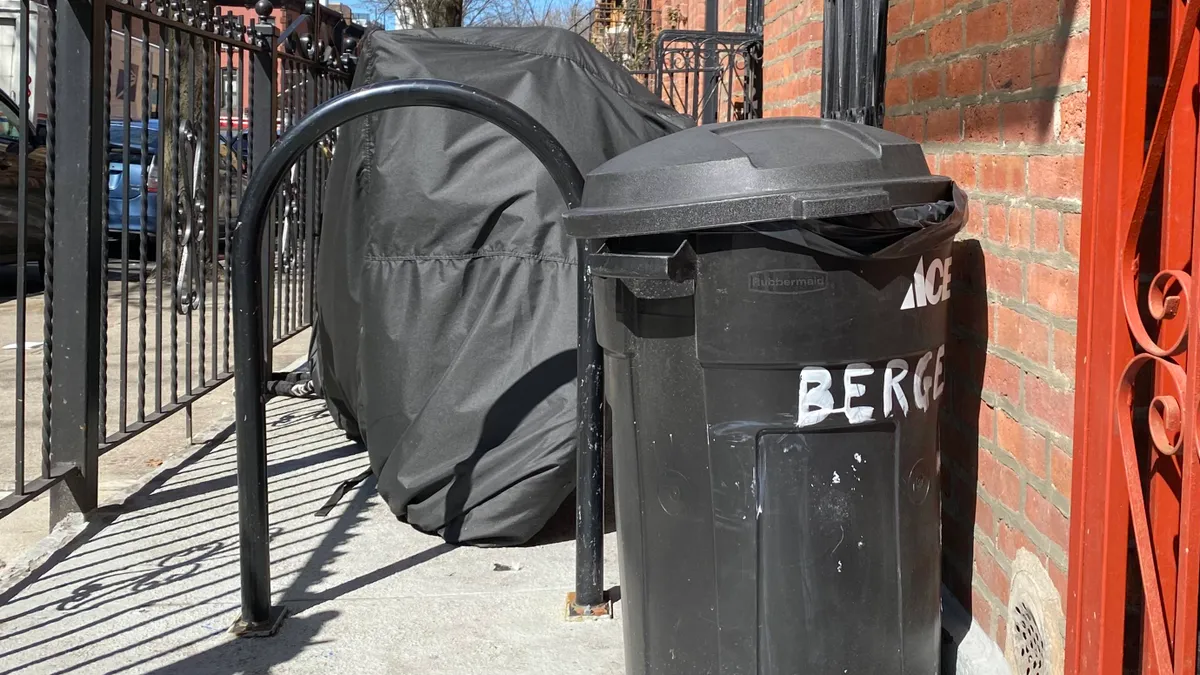 A black lidded trash can sits in front of a building in a fenced-off area. Next to it are a bike rack and covered moped.