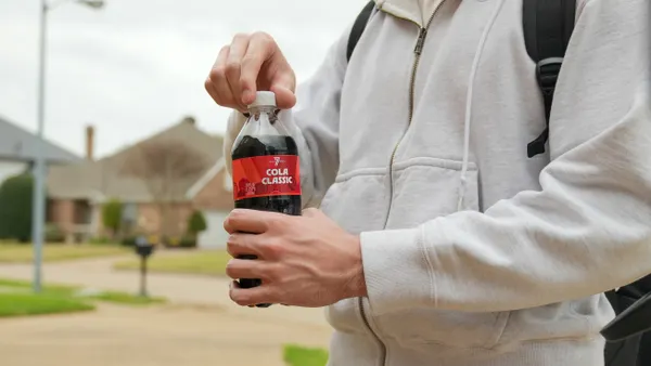 A person is opening a bottle of soda while walking in a neighborhood. The bottle says 7Select cola classic.