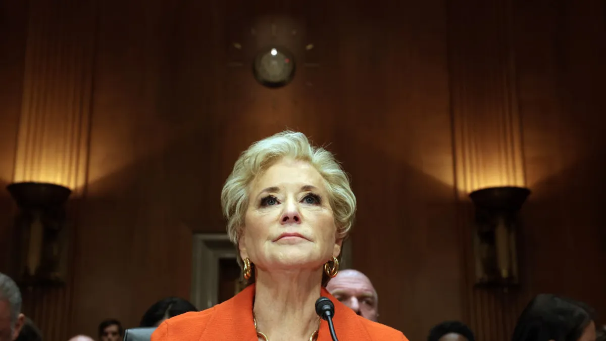 A person is photographed from the front and below. Only the person's head, neck and shoulders can be seen. They are in a room with wood paneling. Others are seated behind the person.