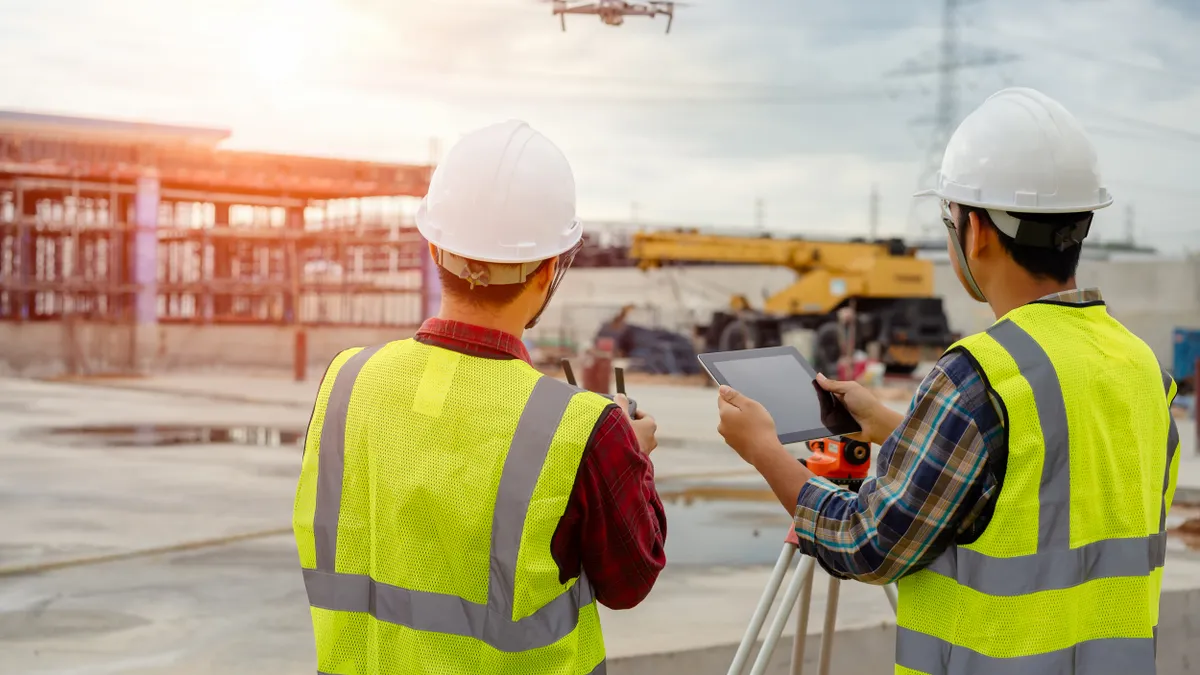 Two construction workers stand as one operates a drone. The drone is in the foreground of the picture, with the two workers who wear hardhats and safety vests.