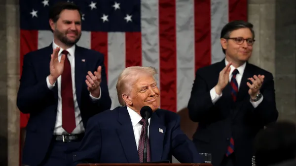 Smiling man in a suit at a podium looking to his left as two men in suits applaud behind him.