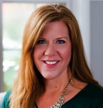 A photo of a smiling woman with red hair, green shirt and wearing a gold necklace.