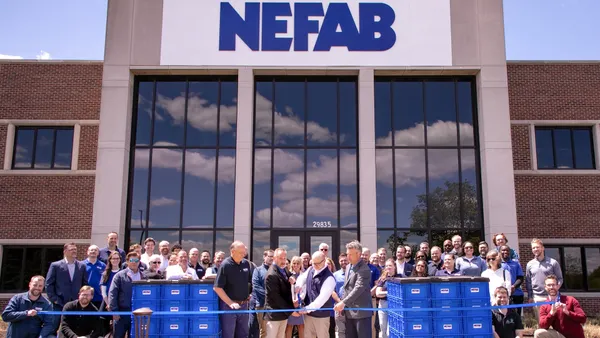 Dozens of people stand behind two people cutting a ribbon in front of a building bearing Nefab's logo.