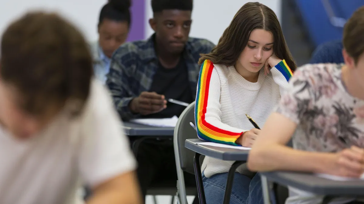 High school students seated at desks, focused on writing during an exam; a girl in a white sweater with rainbow stripes is in the foreground.