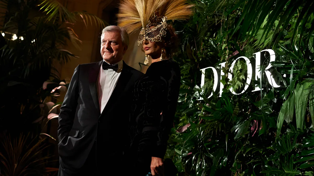 A man in a tuxedo and a woman in a feather mask stand in front of a Dior logo.