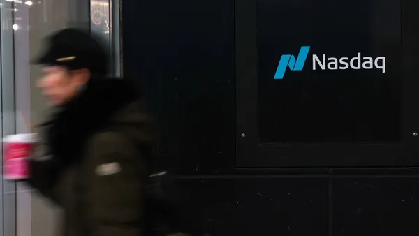 A sign spelling Nasdaq hangs on the side of a building while a pedestrian passes by.