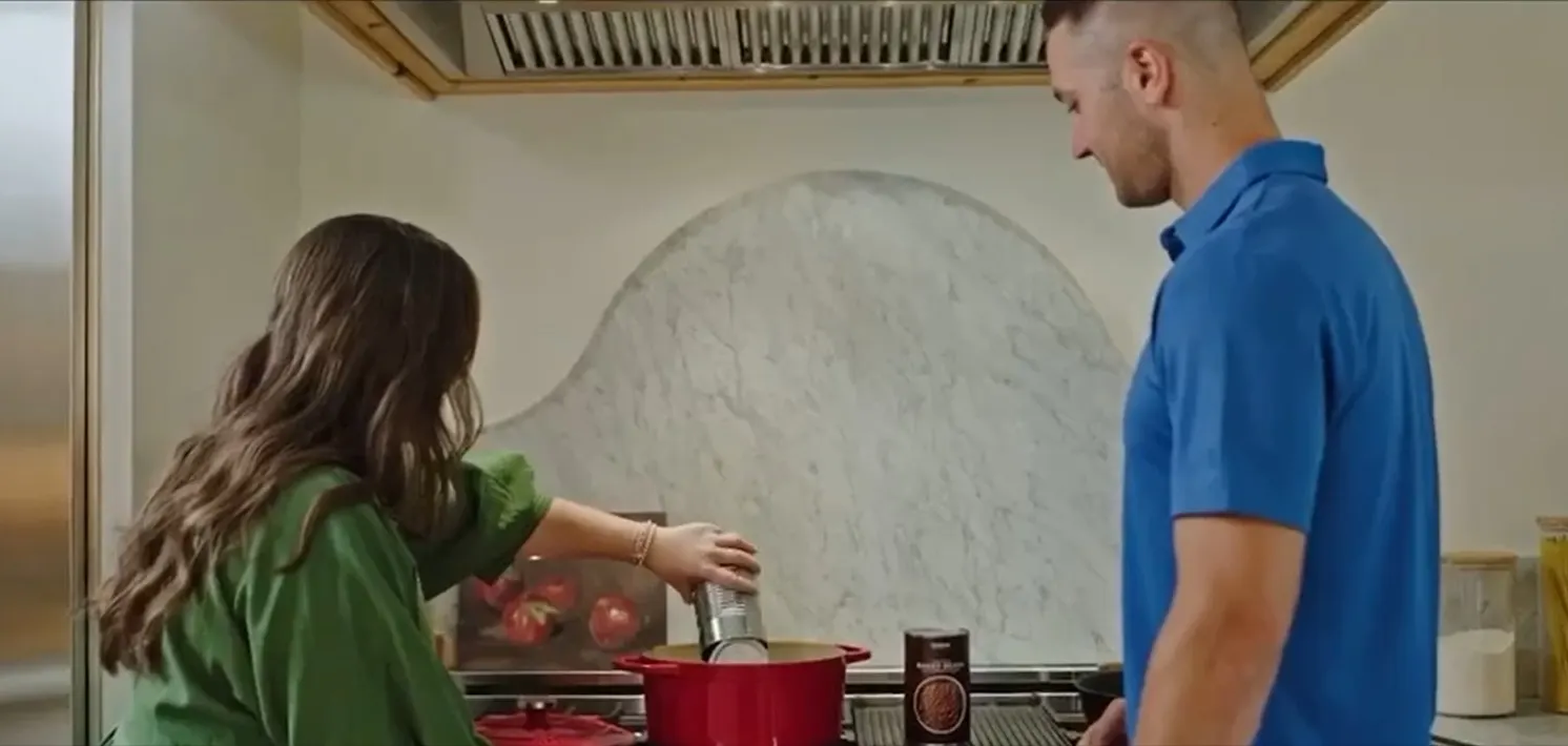 Two people stand next to a stove where one of them is pouring food from a metal can into a pot.