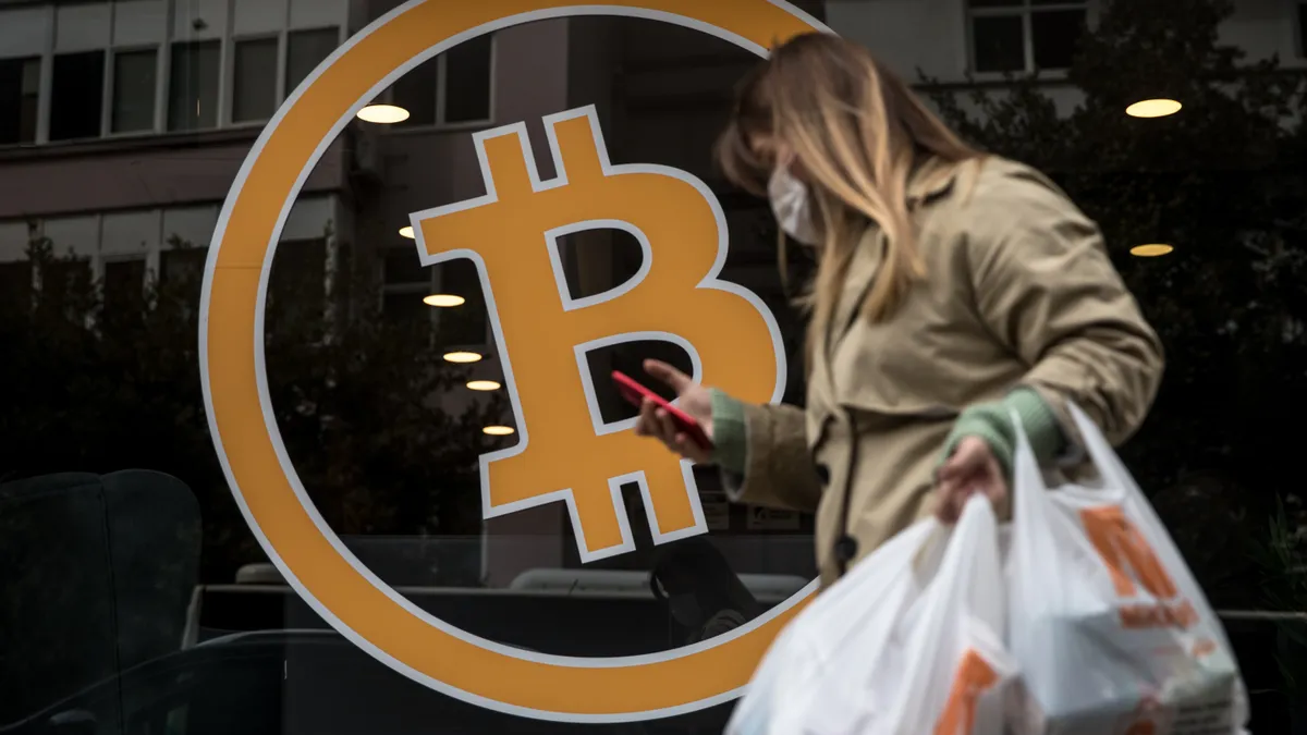 Person with shopping bag passes storefront window with Bitcoin sign