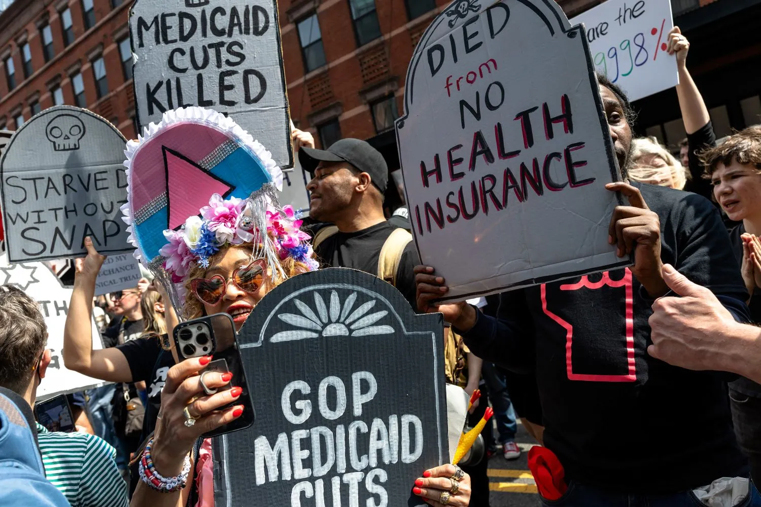 Protestors hold signs shaped like headstones with a woman wearing a flower crown and holding a smartphone in the center.