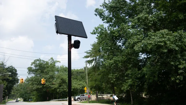 Tall black bar with solar panel and camera on the side of a road with cars in the background and a bike riding by.
