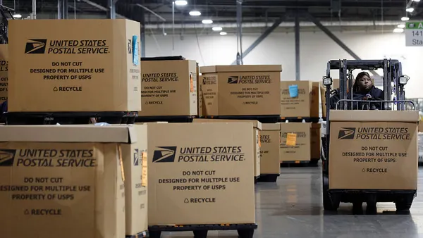 United States Postal Service-branded cardboard boxes stacked in a warehouse, on the right side an employee drives drives a forklift, transporting a box.