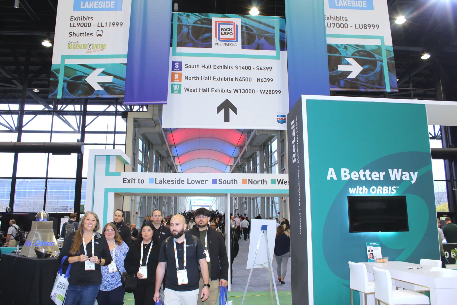 Dozens of people walk through a convention center walkway under signs with directions.