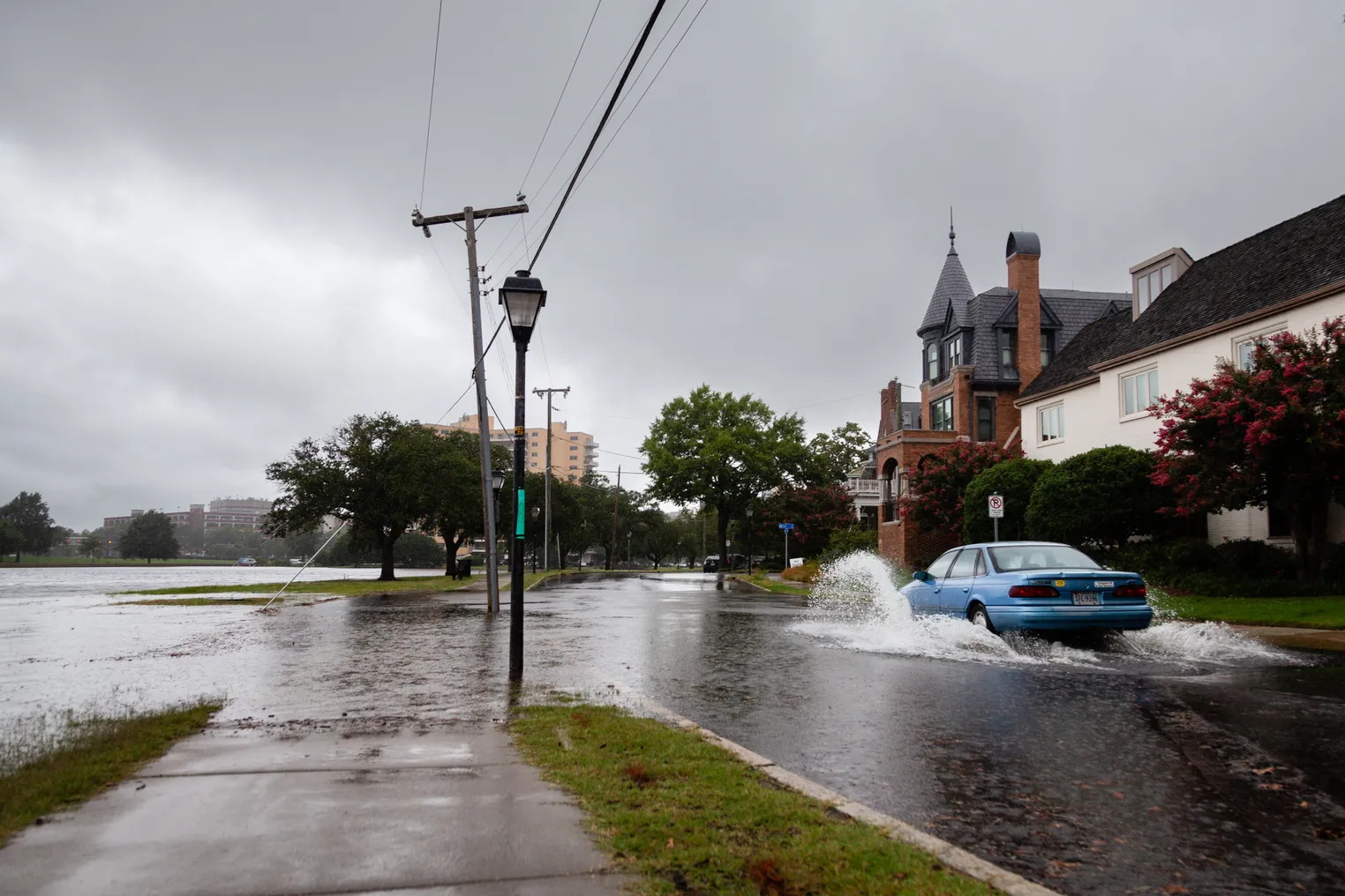 A blue car drives through a flooded street, spraying up water.