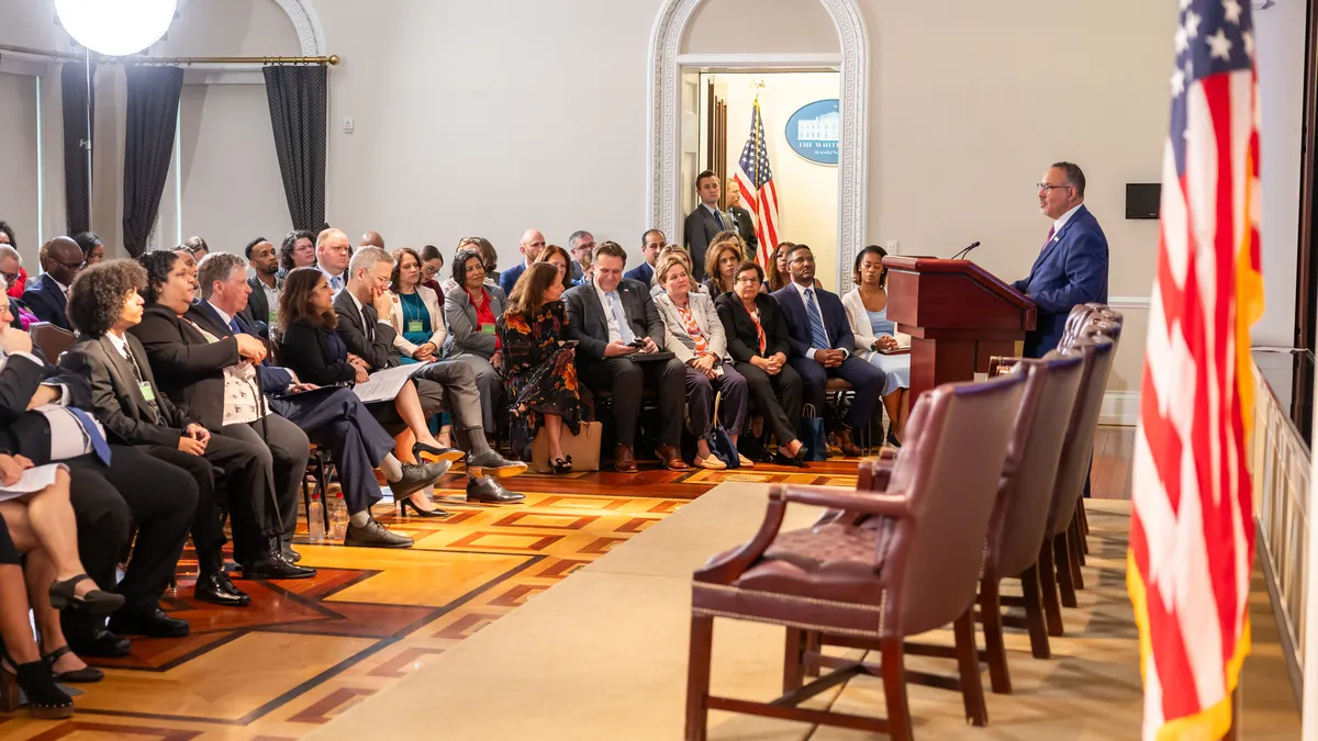 An adult is standing at a podium in the front of a large room with a stage. In the room are rows of seats with people.