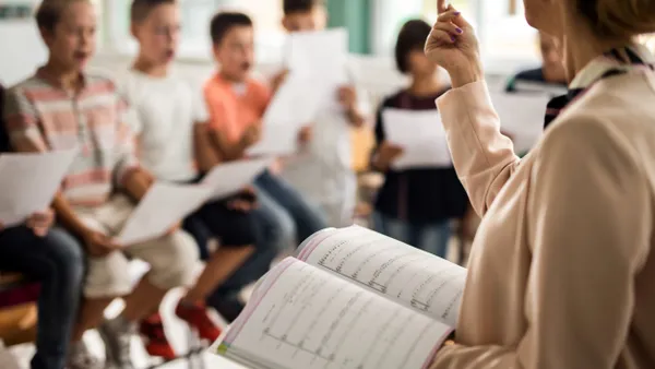 A teacher with sheet music practices with a choir of children in a classroom.