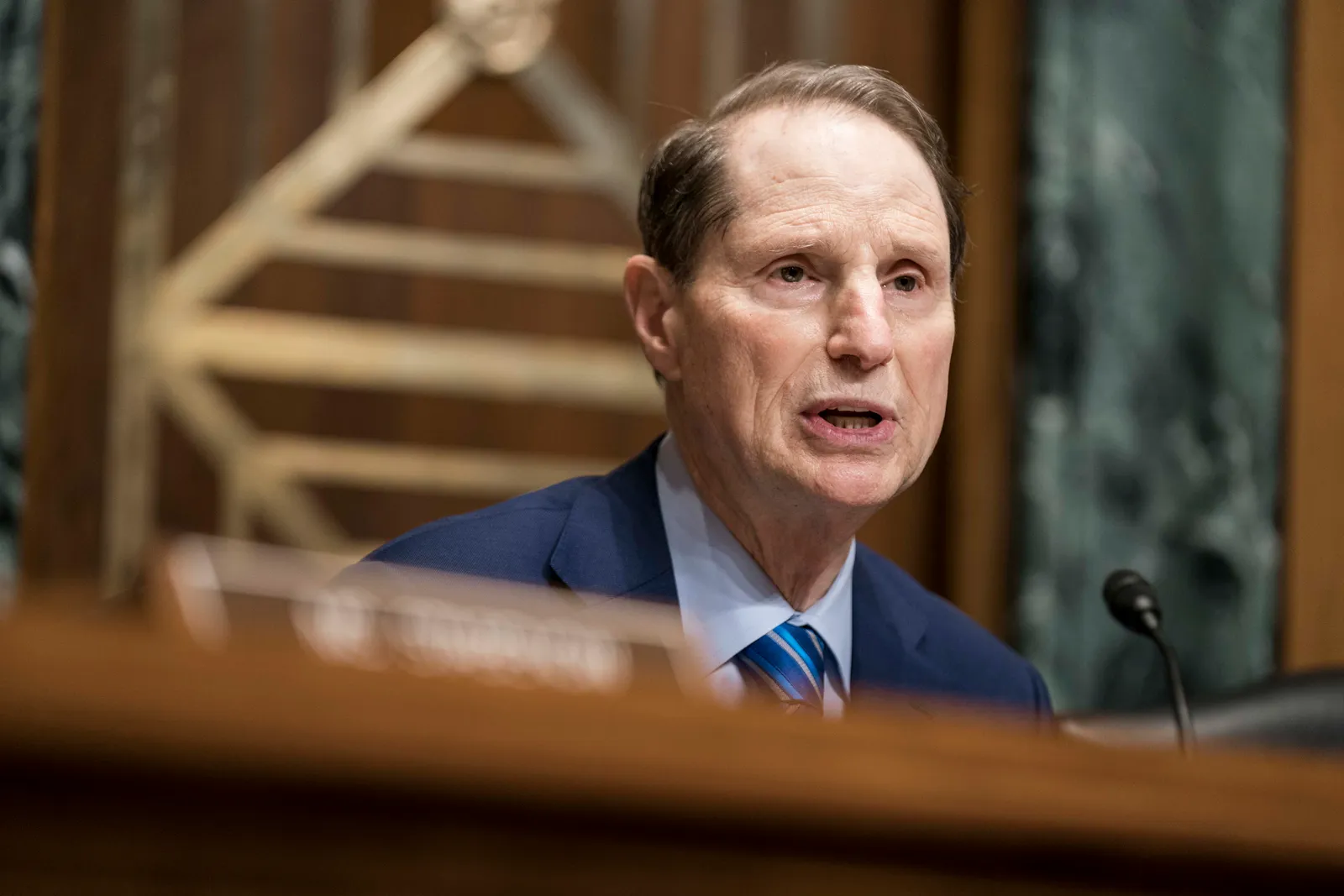 Sen Ron Wyden sits behind a wooden podium with a microphone.