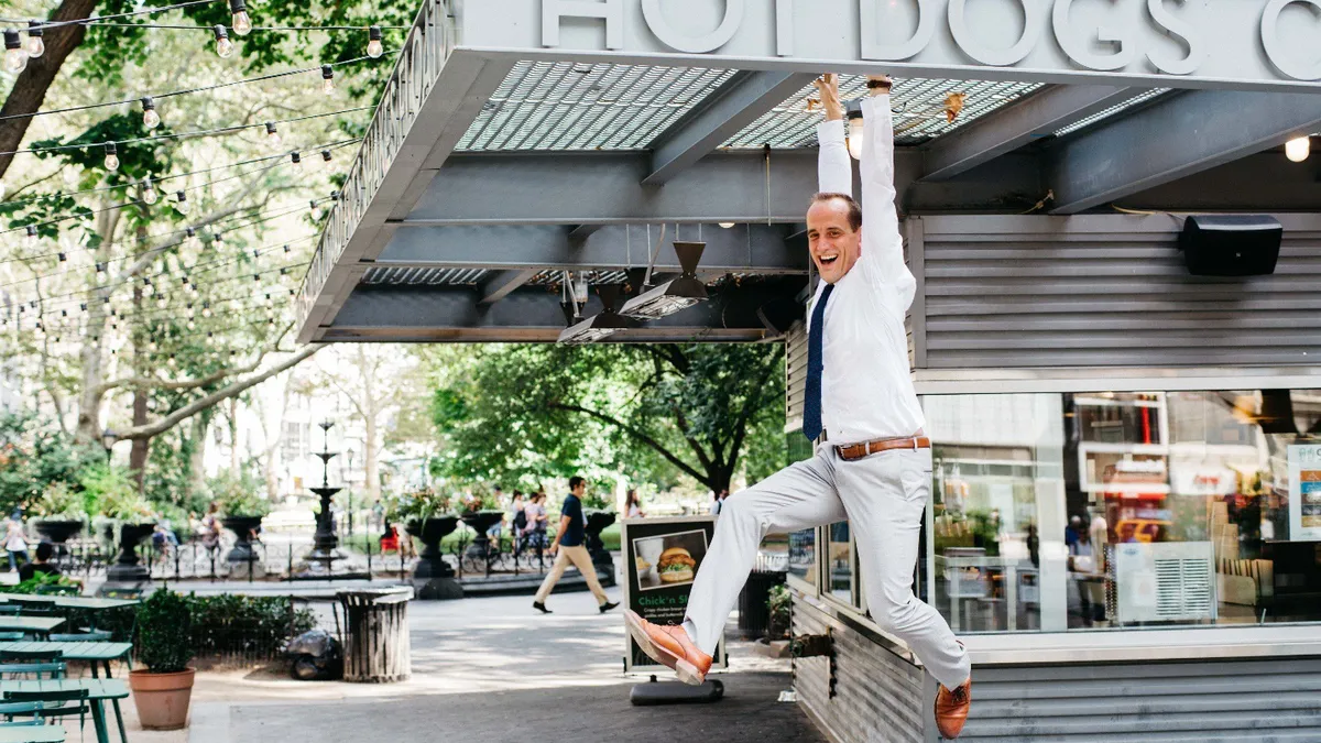 A photograph of Shake Shack CEO Randy Garutti jumping in the air in front of a Shake Shack