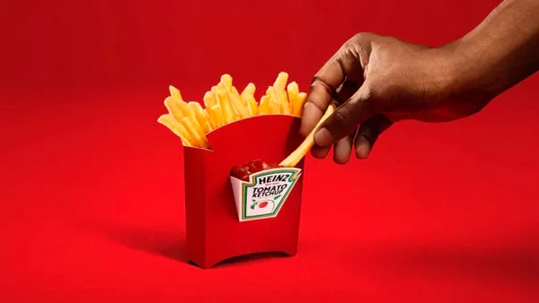 A person's hand dips a french fry into a side compartment that holds ketchup attached to red french fry box.