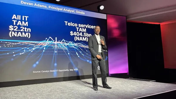 A man in a suit stands on a stage in front of a blue presentation screen. The screen shows his name, "Devan Adams, Principal Analyst, Omdia" and statistics about IT spending.