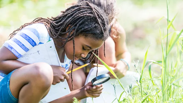 two students are low to the ground looking at vegetation. The student closest to the camera holds a magnifying glass, a notebook and pencil.