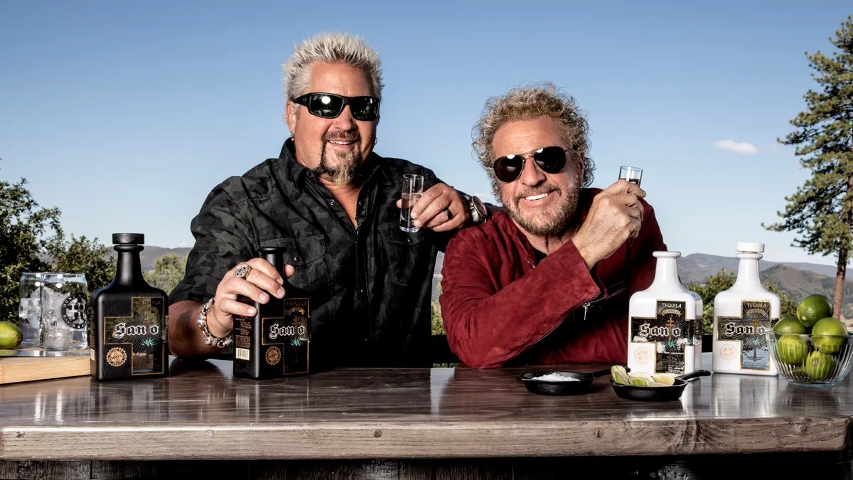 Guy Fieri and Sammy Hagar stand behind a bar raising glasses of spirits.