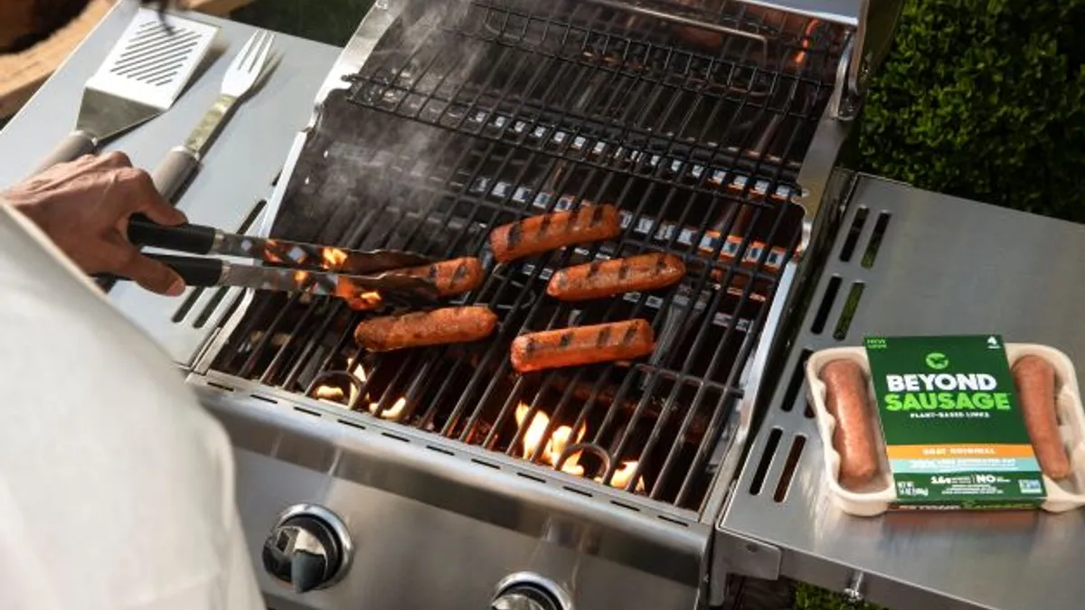 A person cooks Beyond Sausages on an outdoor grill, with the package on the right side.