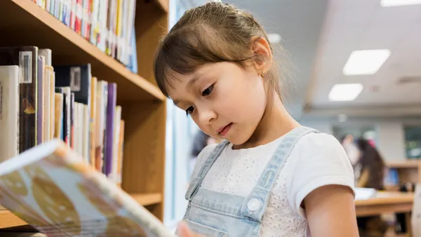 A student is looking down at an open book while standing near a book shelf