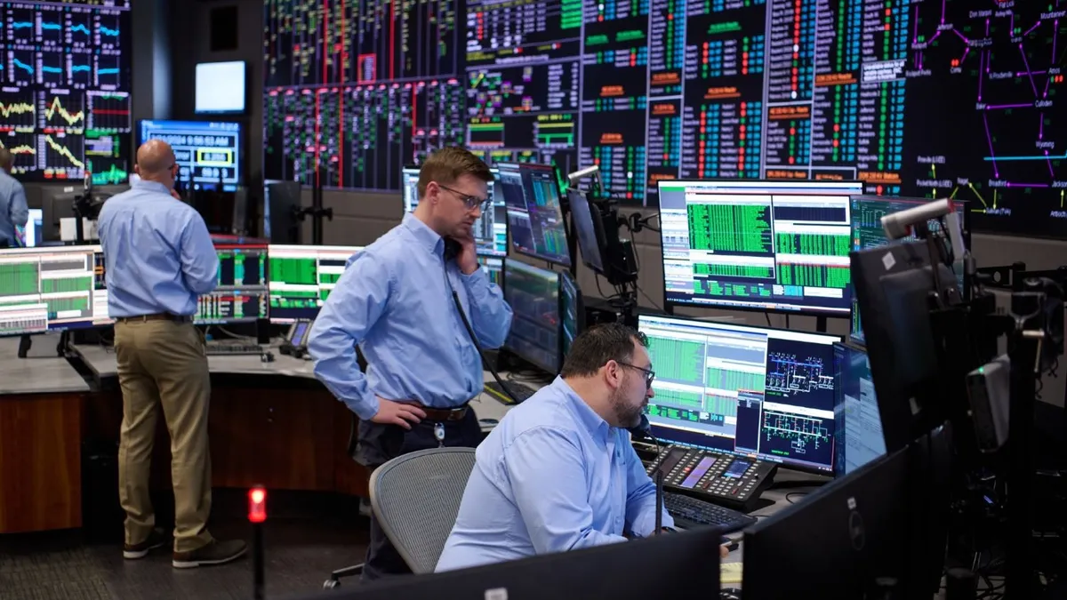 Three men in blue shirts stand at desks with a bank of computer screens.