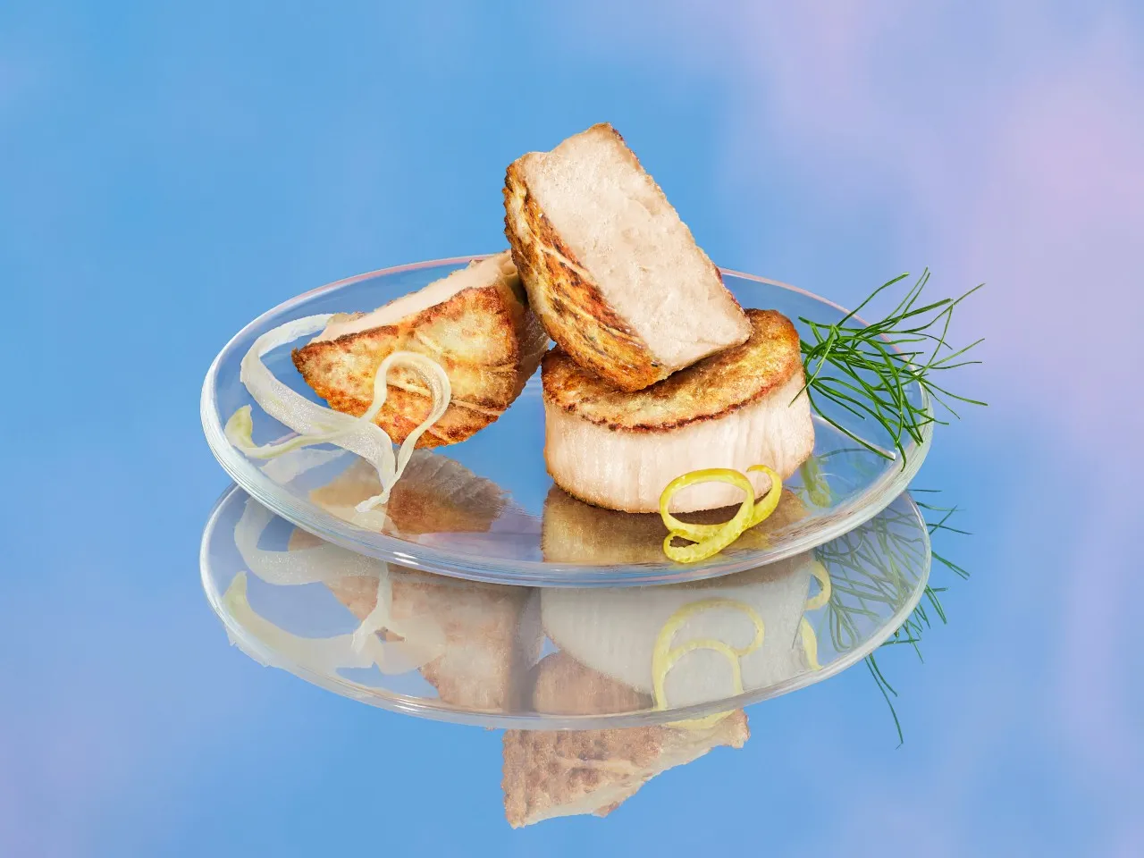 A glass plate with two grilled Air Scallops -- one whole and one cut in half. The plate is garnished with dill and citrus curls, and on a blue surface that looks like the sky.