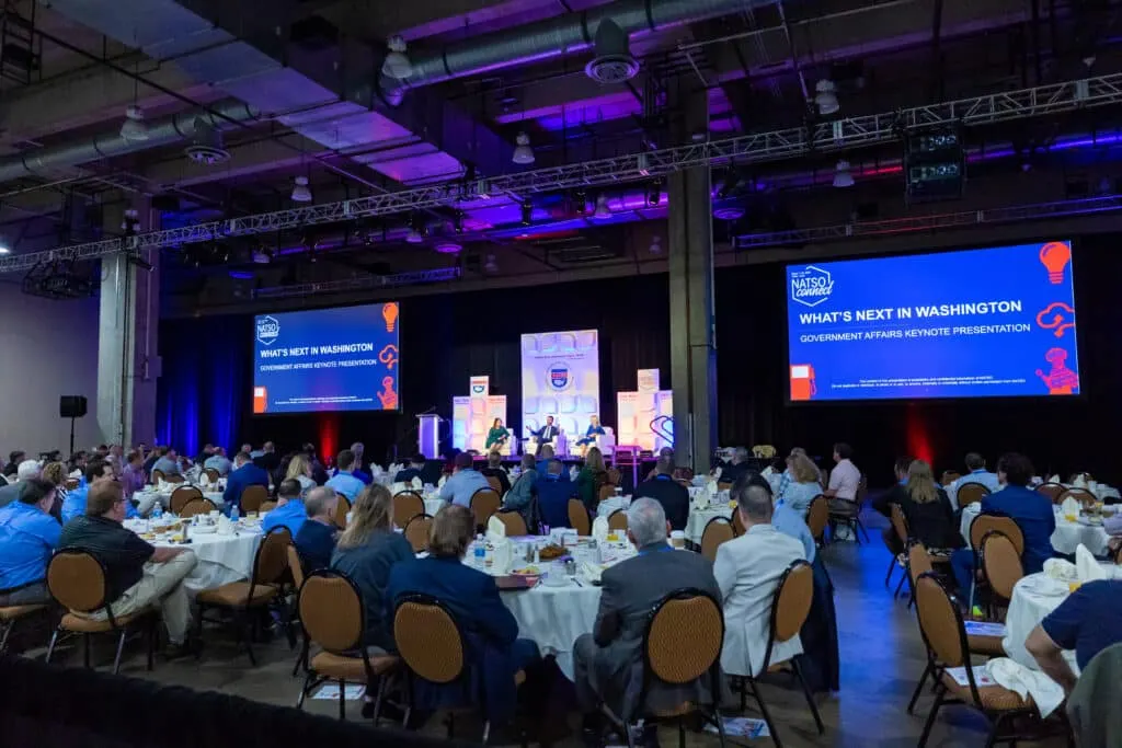 A photo of a number of people sitting at tables in a room listening to a presentation. Two screens at the front of the room say "what's next in Washington."