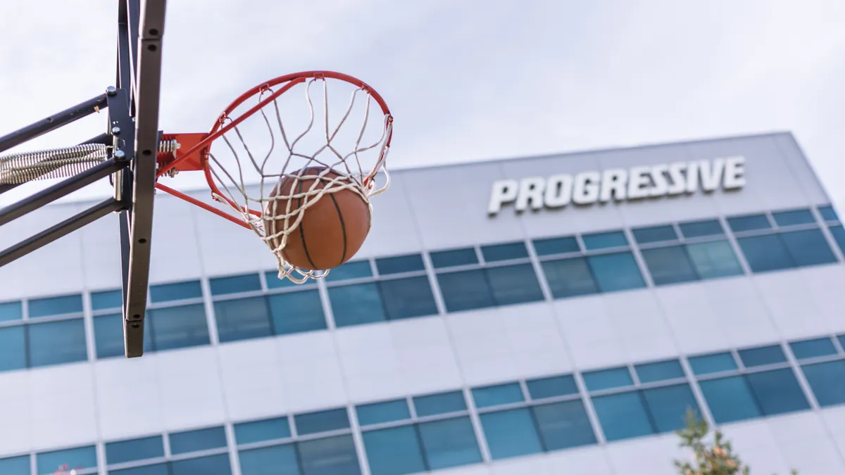 A basketball hoop is seen in the foreground, with an office building featuring the Progressive logo in the background.