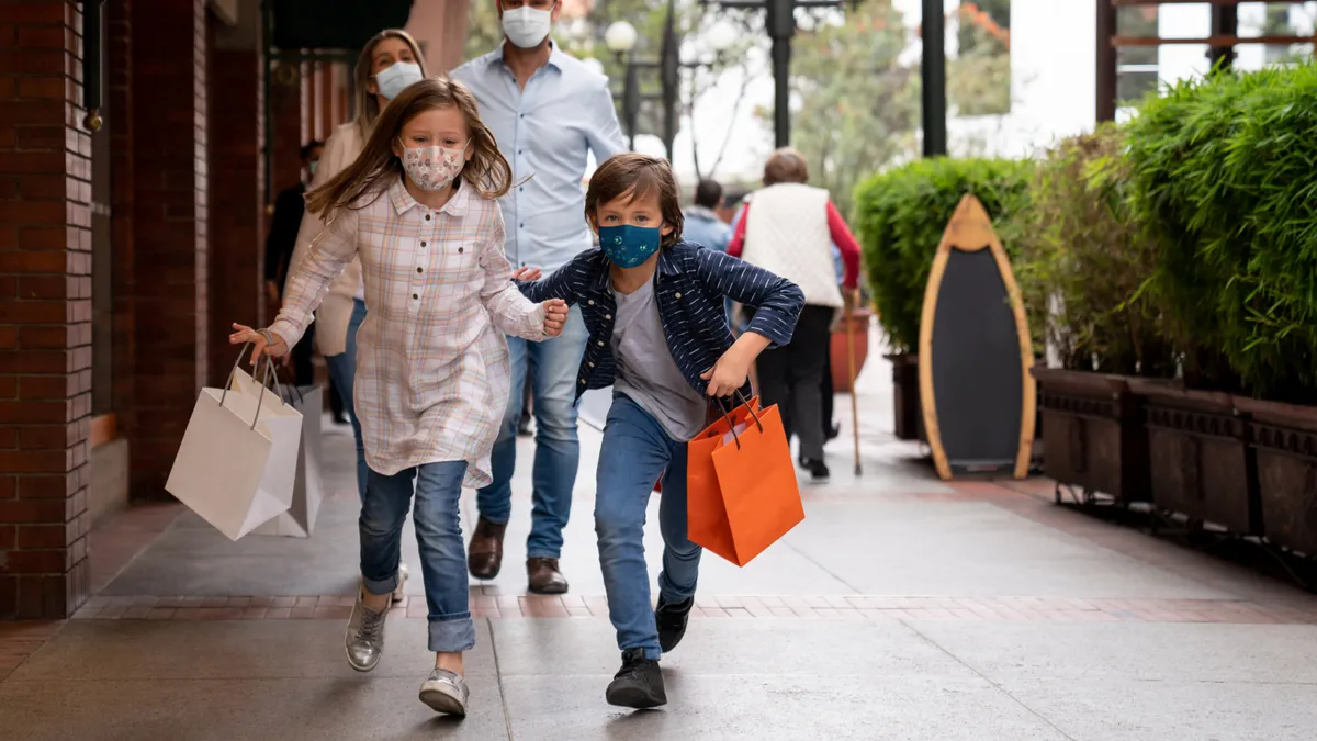 Two kids running with shopping bags in front of two adults outside on a sidewalk near a building.